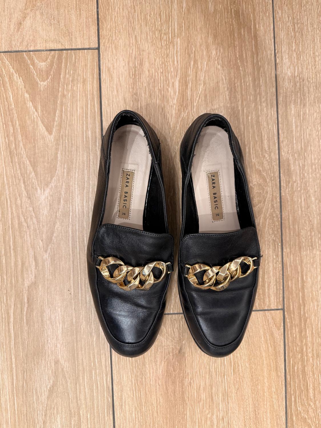 Black leather shoes with a gold chain on the top, on a light wooden floor. Inside label: "ZARA BASIC".