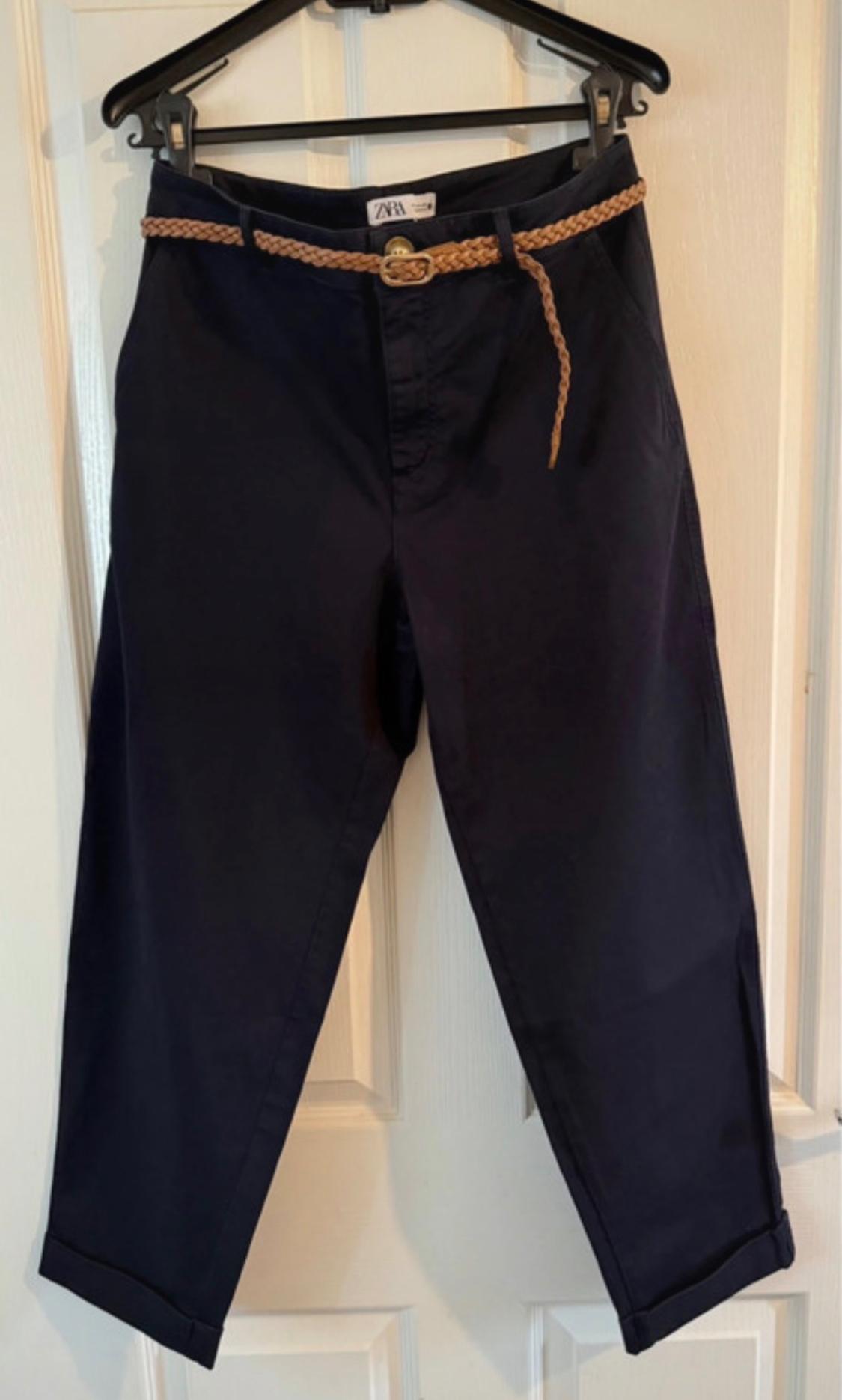 Navy blue trousers hanging on a black hanger, with a light brown braided belt. Visible label: "ZARA". White door background.