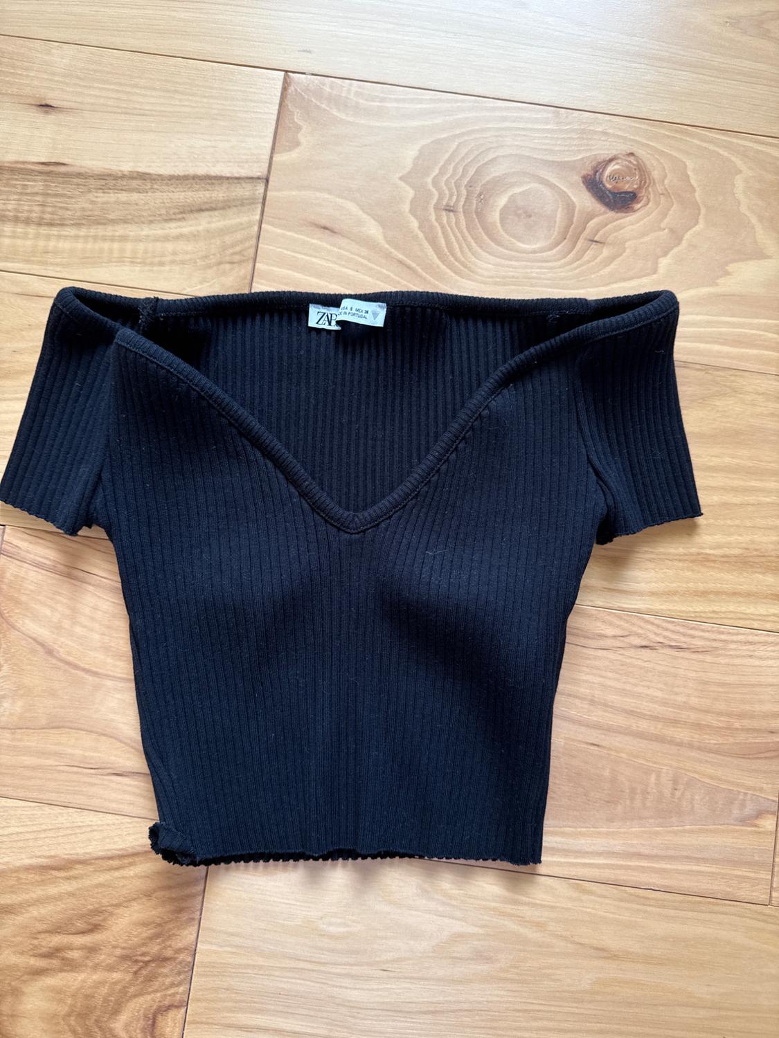 Black ribbed crop top with a V-neck, visible label with the text "ZARA", placed on a light wooden floor.