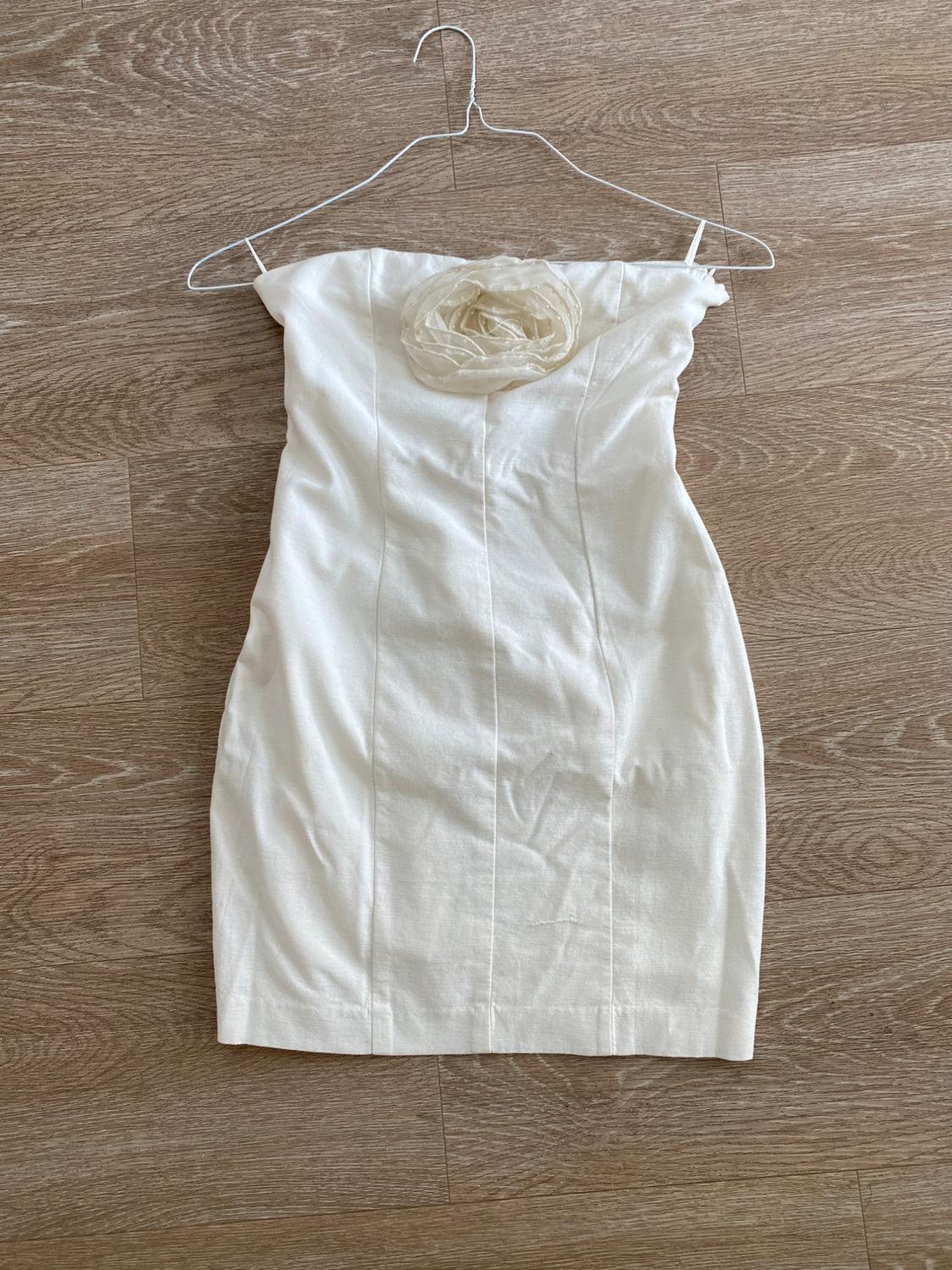 Strapless white dress with a floral detail on the bust, hanging on a hanger over a light wood floor.