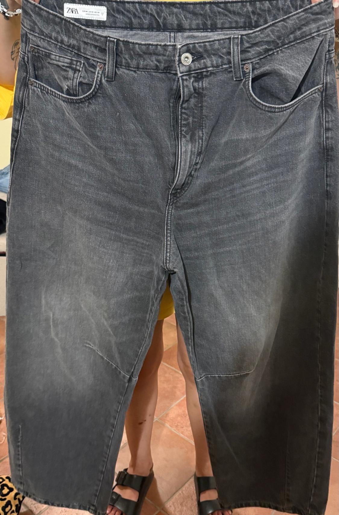 A person holds a pair of oversized grey jeans in front of their body. Their legs can be seen wearing black sandals, along with the label bearing the text ZARA.