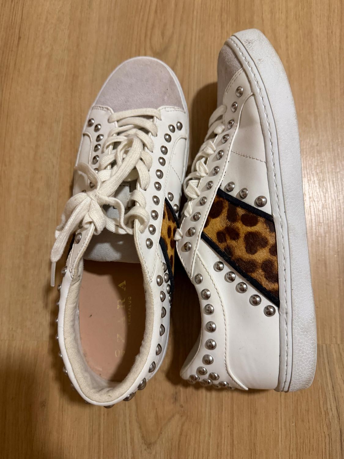 White trainers with silver studs and animal print details on the sides, on a light wooden floor.