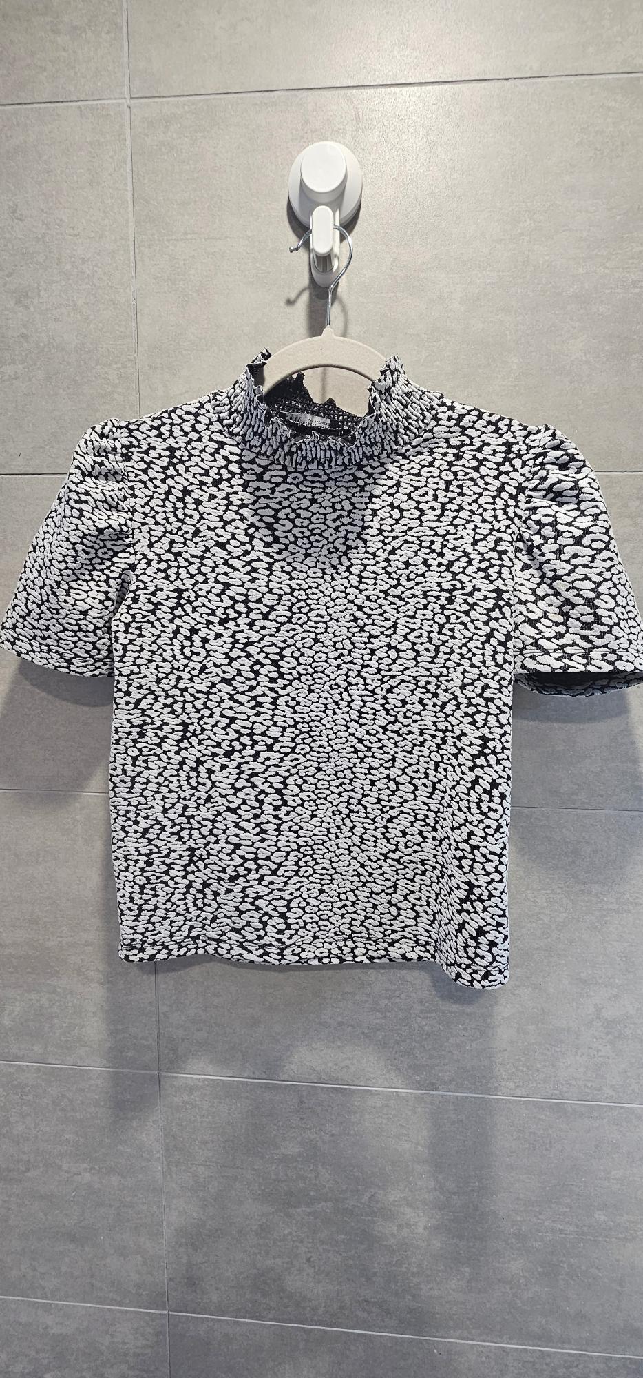 Black and white printed short-sleeved top hanging on a hanger against a grey tiled wall.