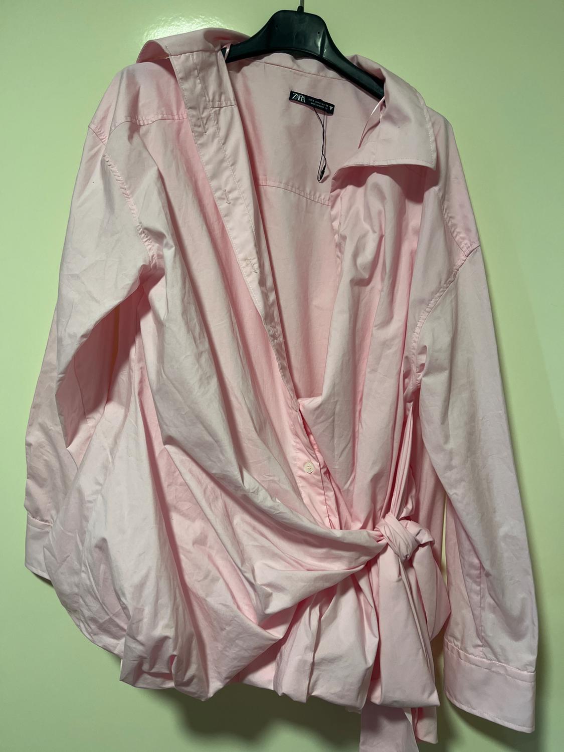 Light pink shirt with a wrap design and a tie waist, hanging on a black hanger against a light green background.