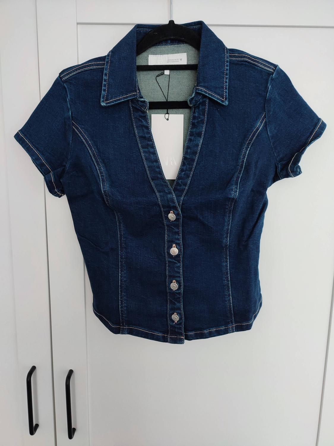 Dark blue denim short-sleeved top with a V-neck and front buttons, hanging on a black hanger in front of a white wardrobe.