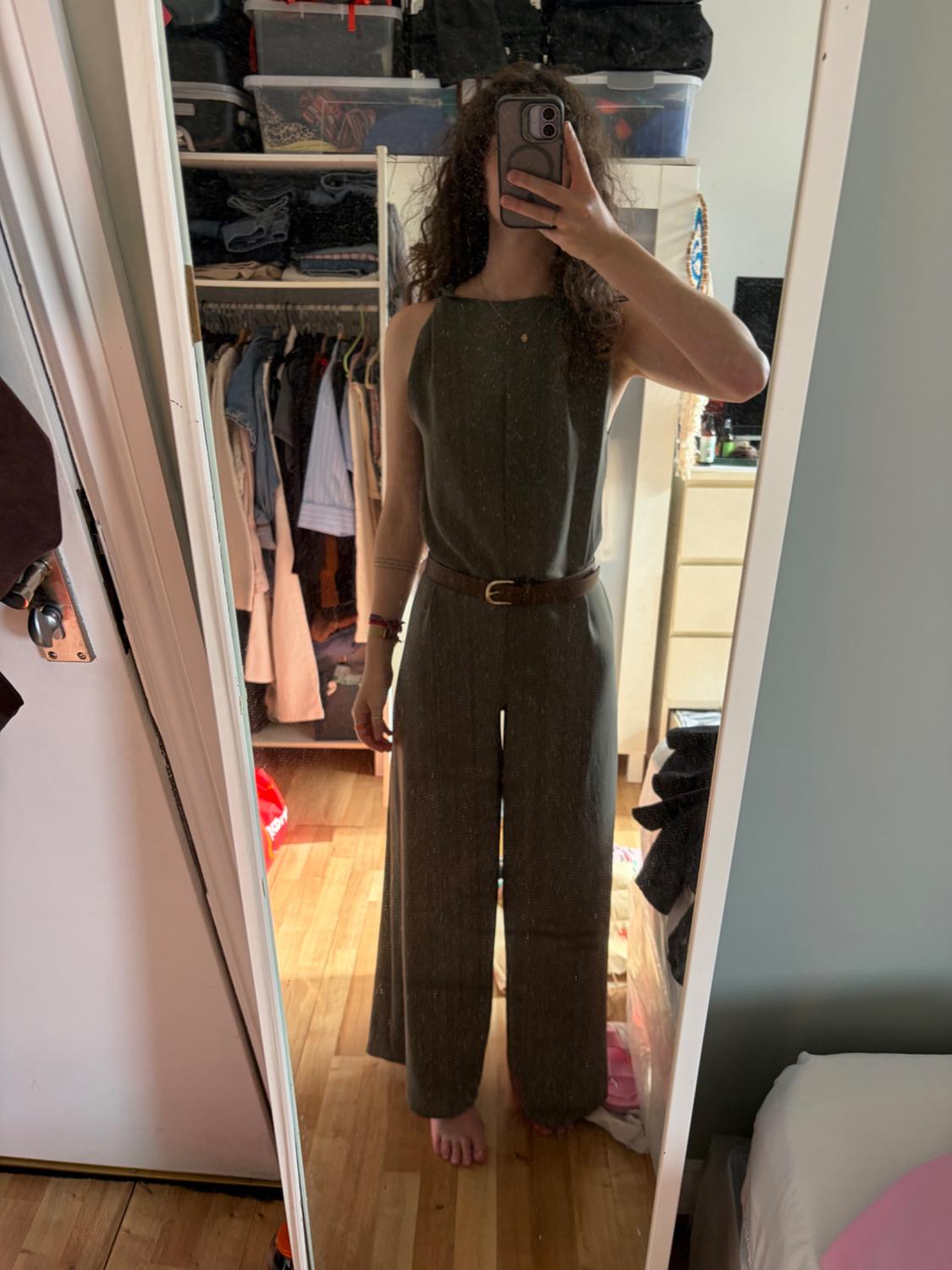 Woman taking a selfie in her bedroom mirror. She is wearing a sleeveless olive green jumpsuit and a brown belt. Casual style.