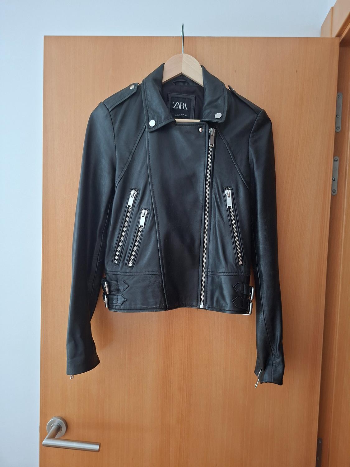 Black biker jacket with zips and metal details, hanging on a wooden door. Inner label with the text ZARA.