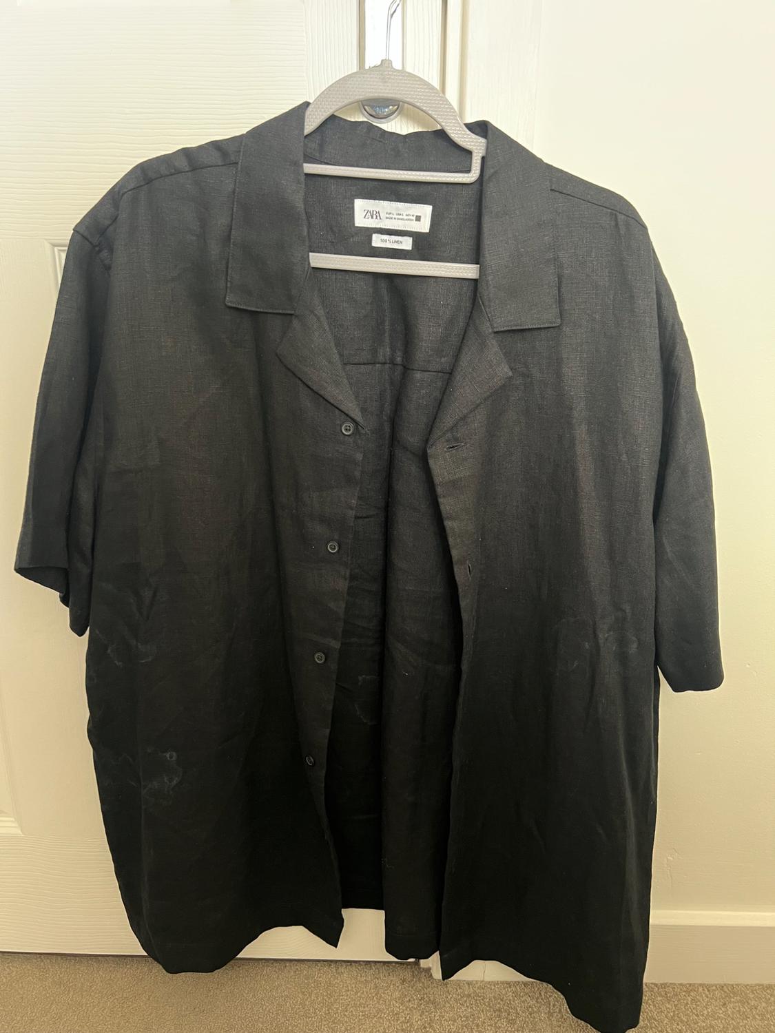 Black short-sleeved shirt with a lapel collar hanging on a hanger over a white door. Label with the text ZARA and 100% LINEN.