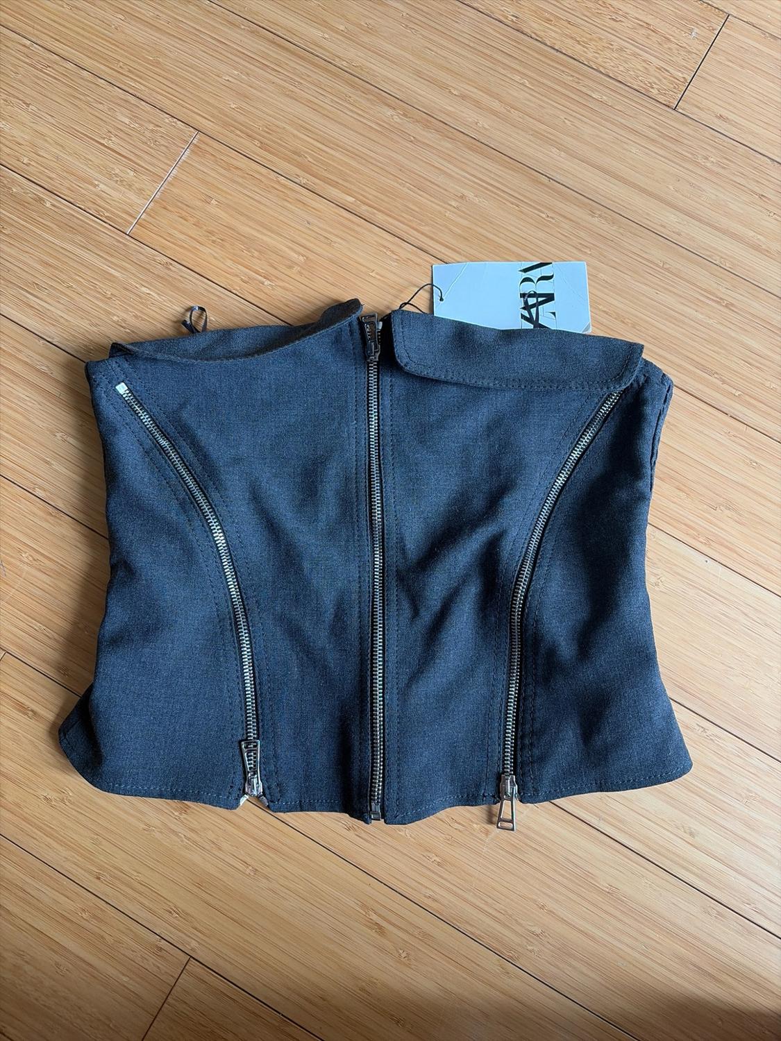 Dark gray strapless top with a folded hem and three front zippers on a wooden floor. Tag with text Zara.
