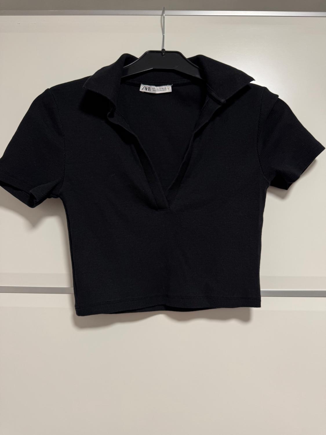 Black short-sleeved open-collar T-shirt, hanging on a black hanger against a white background. Visible label on the collar.