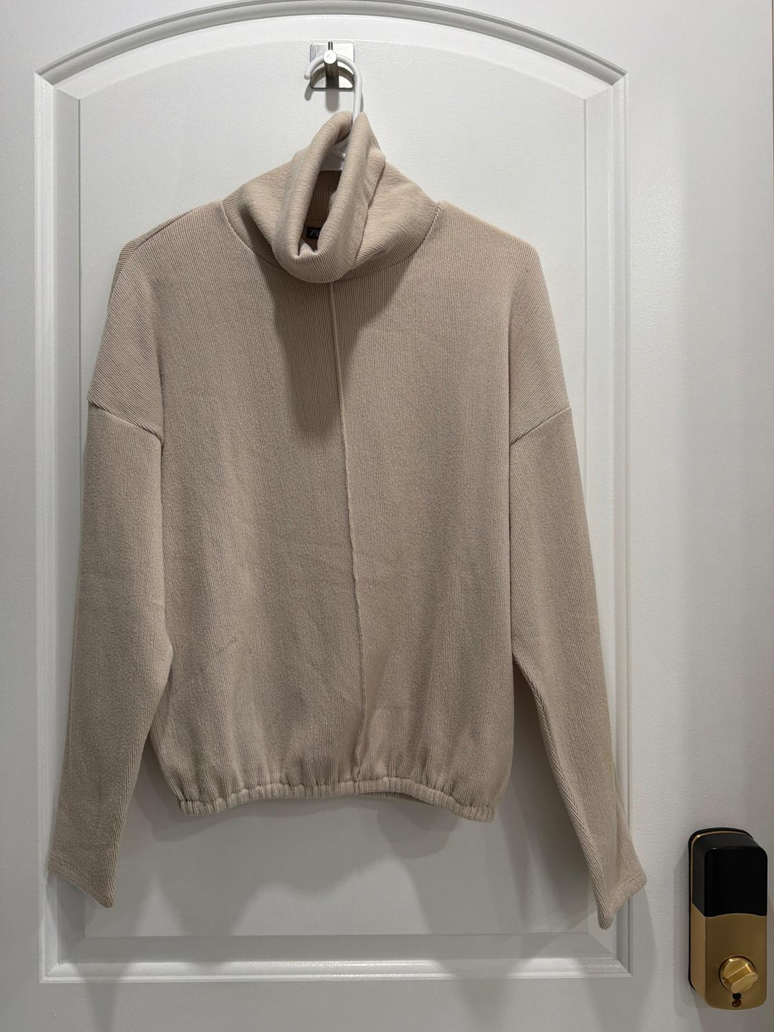 Beige turtleneck sweater hanging on a white door. The door has a gold and black doorknob.