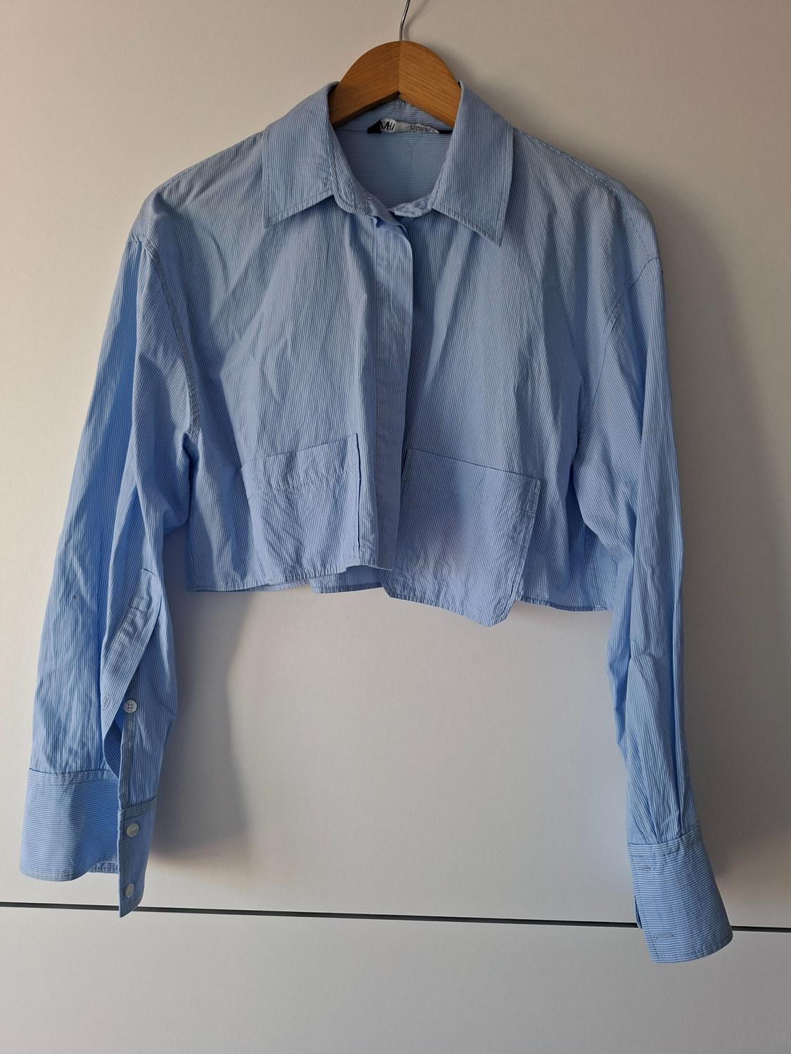 Light blue striped cropped shirt with front pockets, hanging on a wooden hanger against a white background.