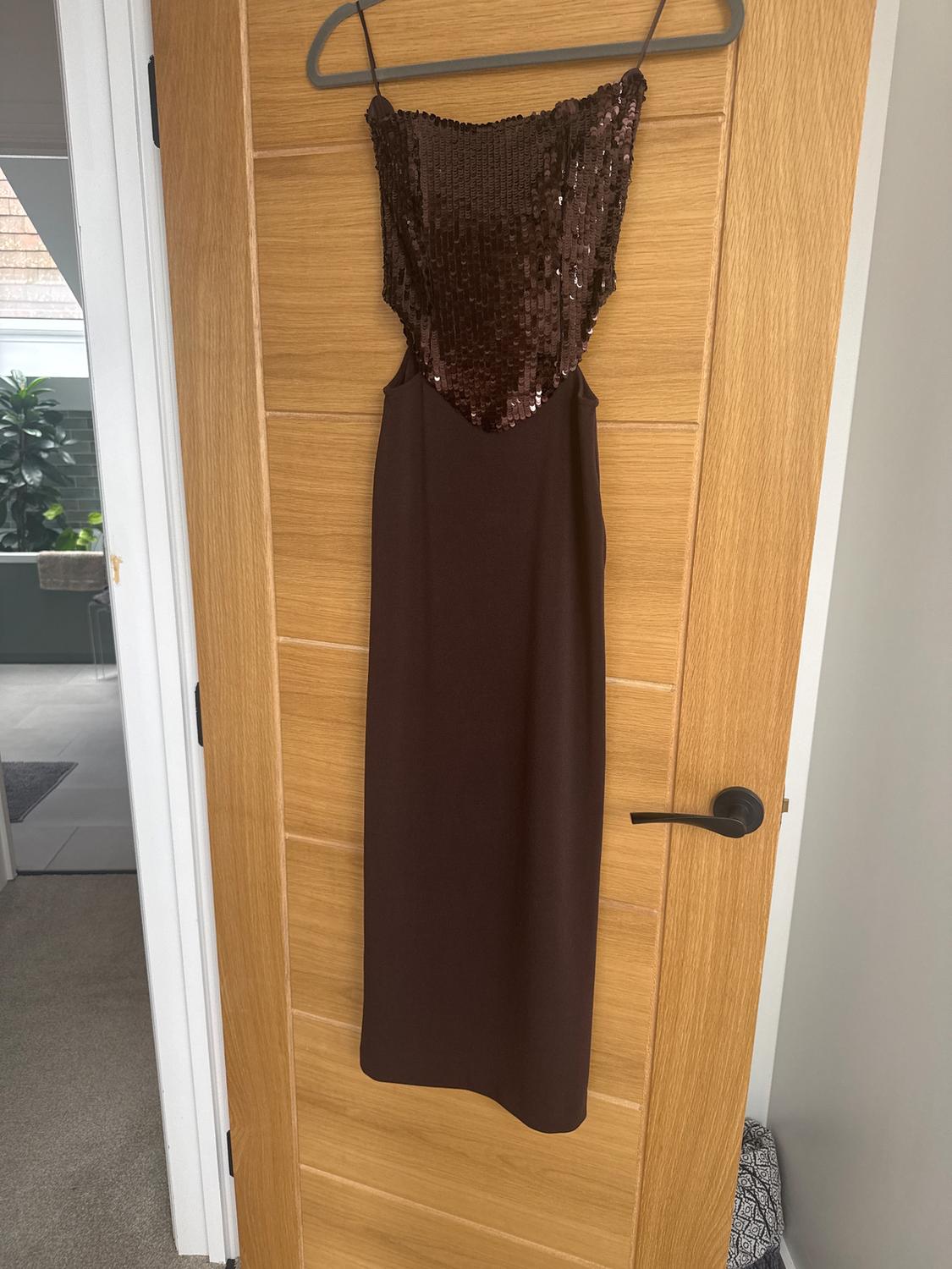 Brown dress with sequins on the top, hanging on a light wooden door. Modern indoor setting.