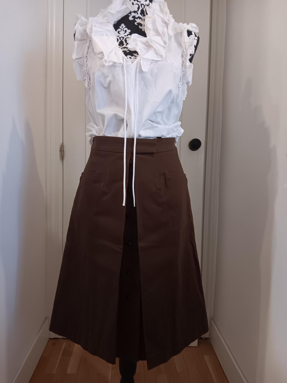 Mannequin in a white ruffled blouse and brown skirt with front buttons. White wall and wooden floor background.