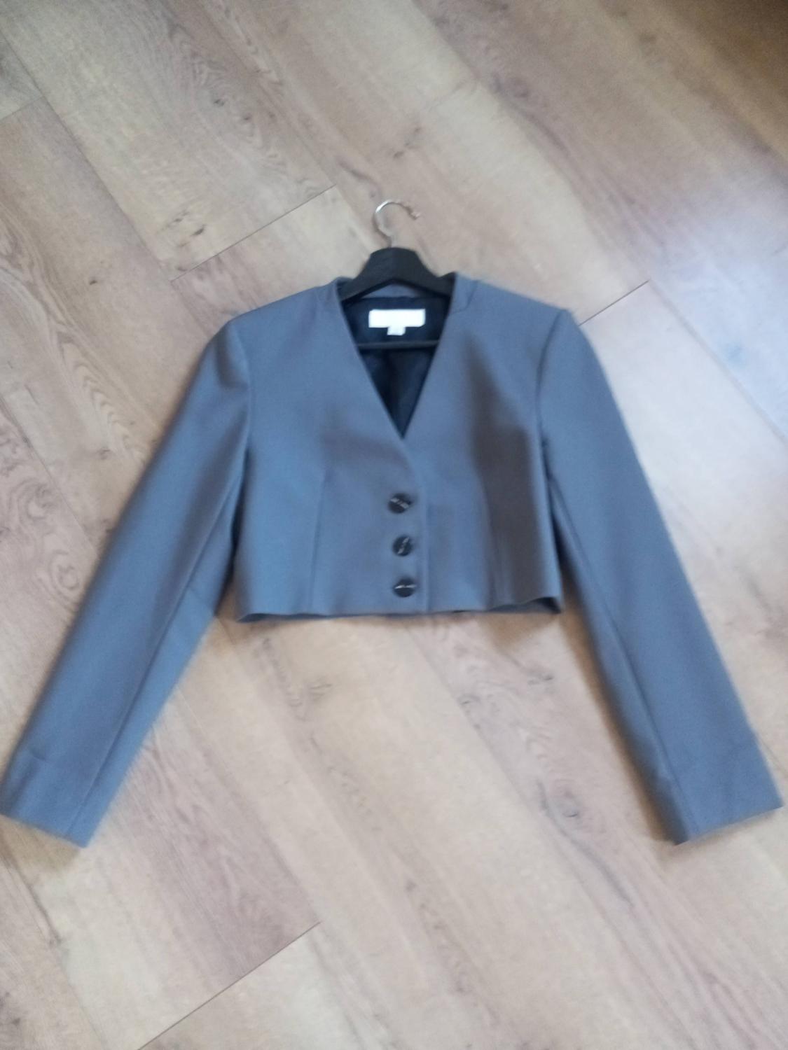 Short grey jacket with three buttons, hanging on a hanger over a light wooden floor.