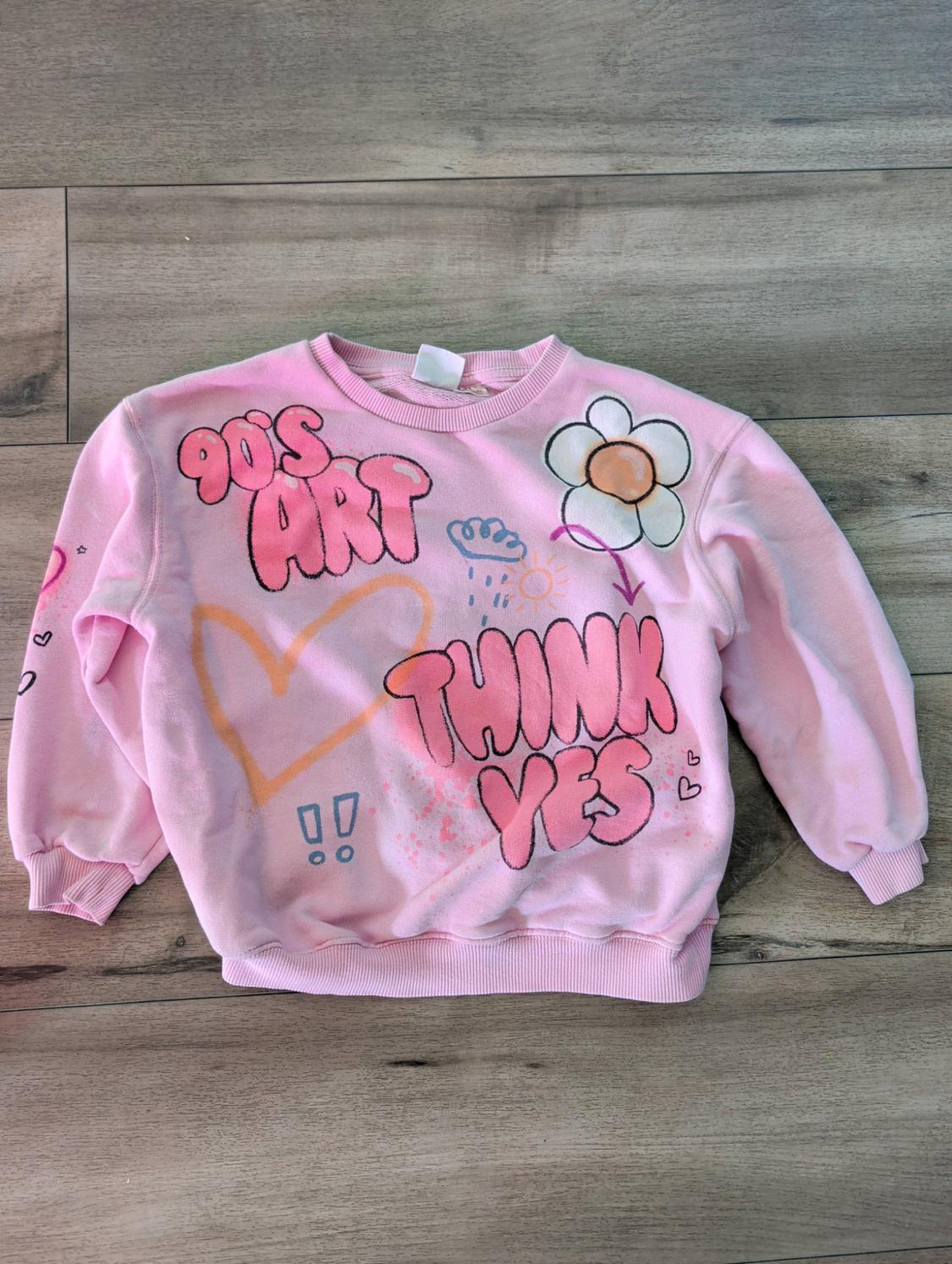 Pink sweatshirt on a wooden floor with flower, heart and cloud drawings, alongside the texts “90'S ART” and “THINK YES”.