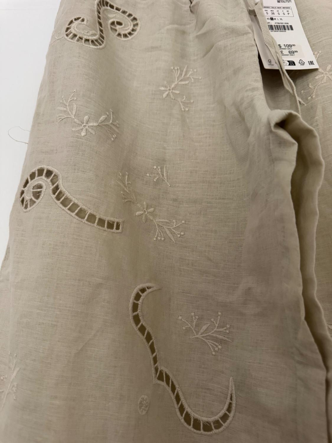 Detail of beige garment with floral embroidery and openwork. Label with text: S 26 S S P XS S M L XL $ 109.00 £ 69.99.