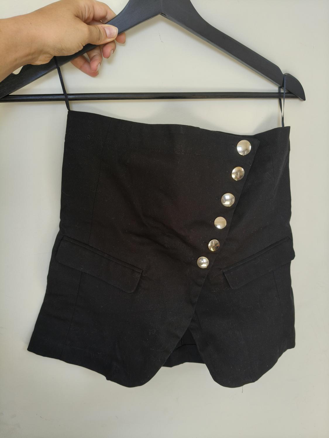 A hand holds a hanger with a black strapless top. It features an asymmetric fastening with silver buttons and flap pockets.