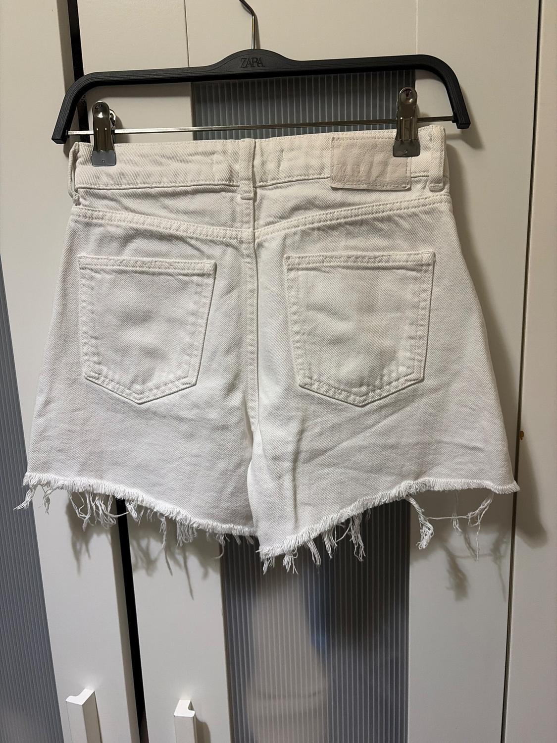 White denim shorts with frayed hem hanging on a black Zara hanger, in front of a white wardrobe.
