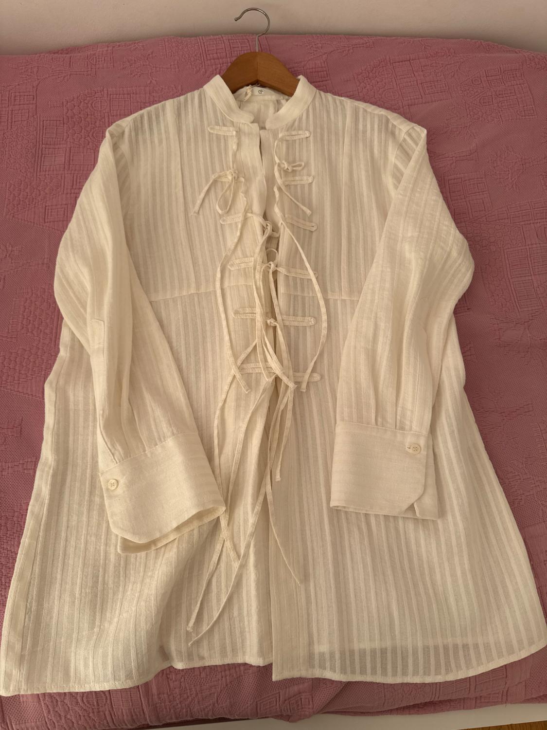 White shirt with vertical stripes, mandarin collar and front ties. Hanging on a wooden hanger against a textured pink background.