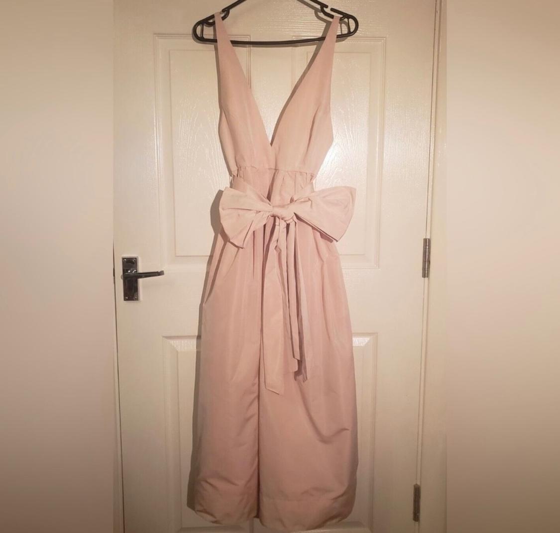 Light pink jumpsuit with V-neck and large bow at the waist, hanging on a hanger over a white door.