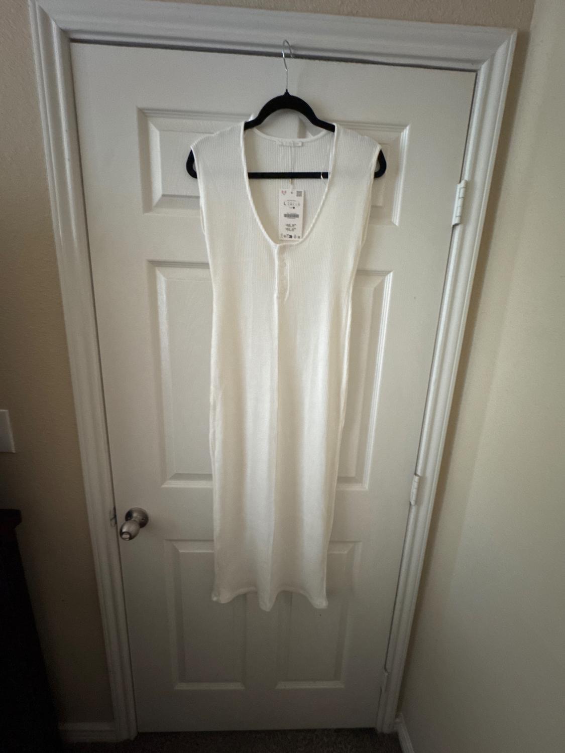 Sleeveless white dress hanging on a white door. Visible tag on the neck. Lit indoor environment.