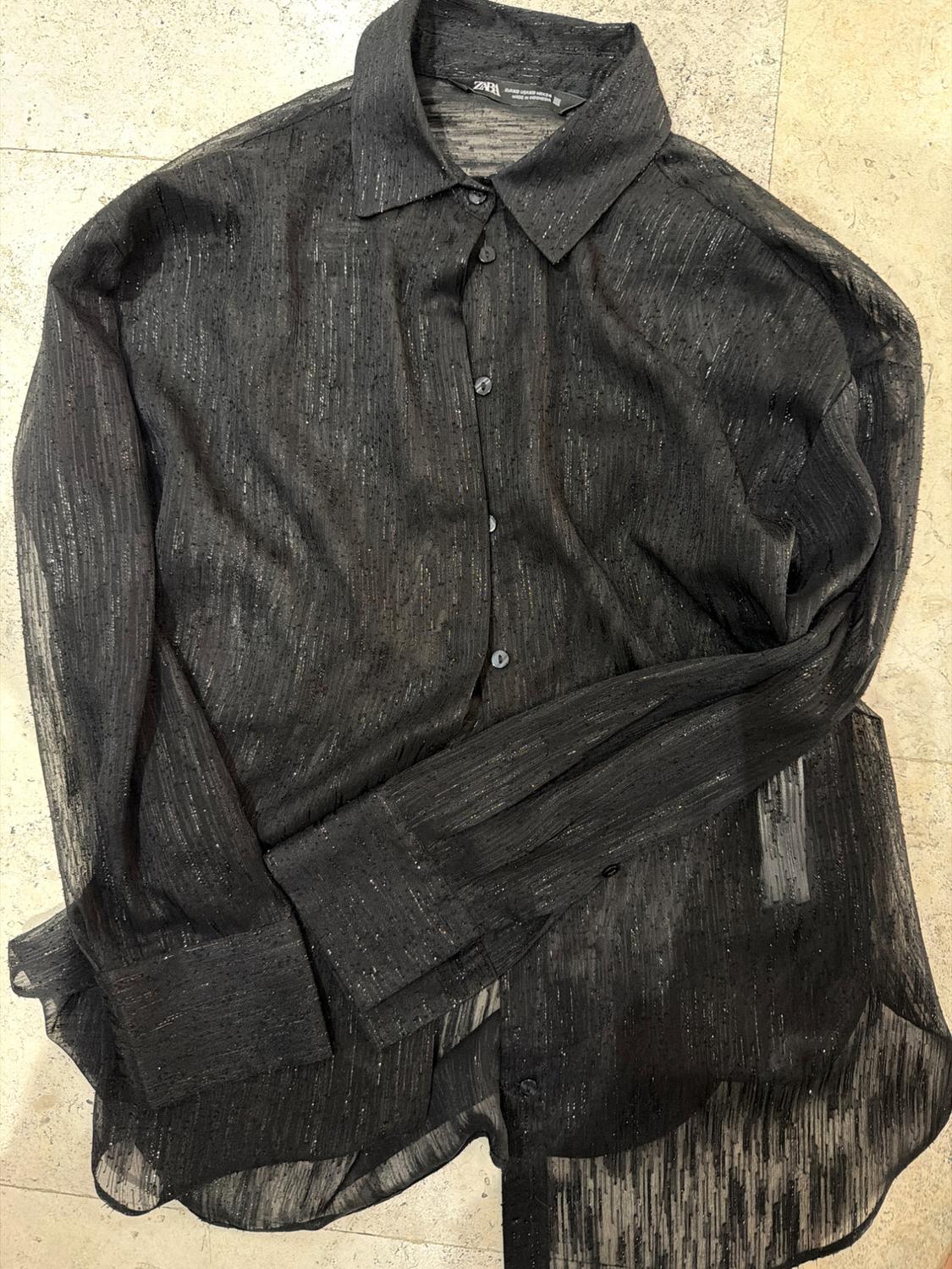 Black semi-sheer long-sleeved shirt with shimmering threads, laid out on a light stone floor. Label with the text ZARA.