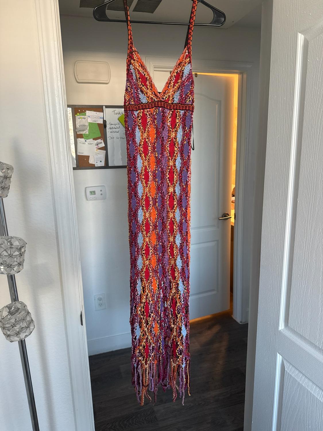 Multicolored crochet maxi dress with diamond patterns and fringe hanging on a door. In the background, a board with the word "Tasks".