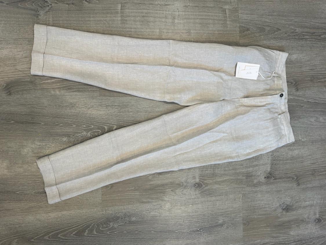 Light beige trousers with a front crease and hem, laid out on a wooden floor. They have a dark button and a label on the waistband.