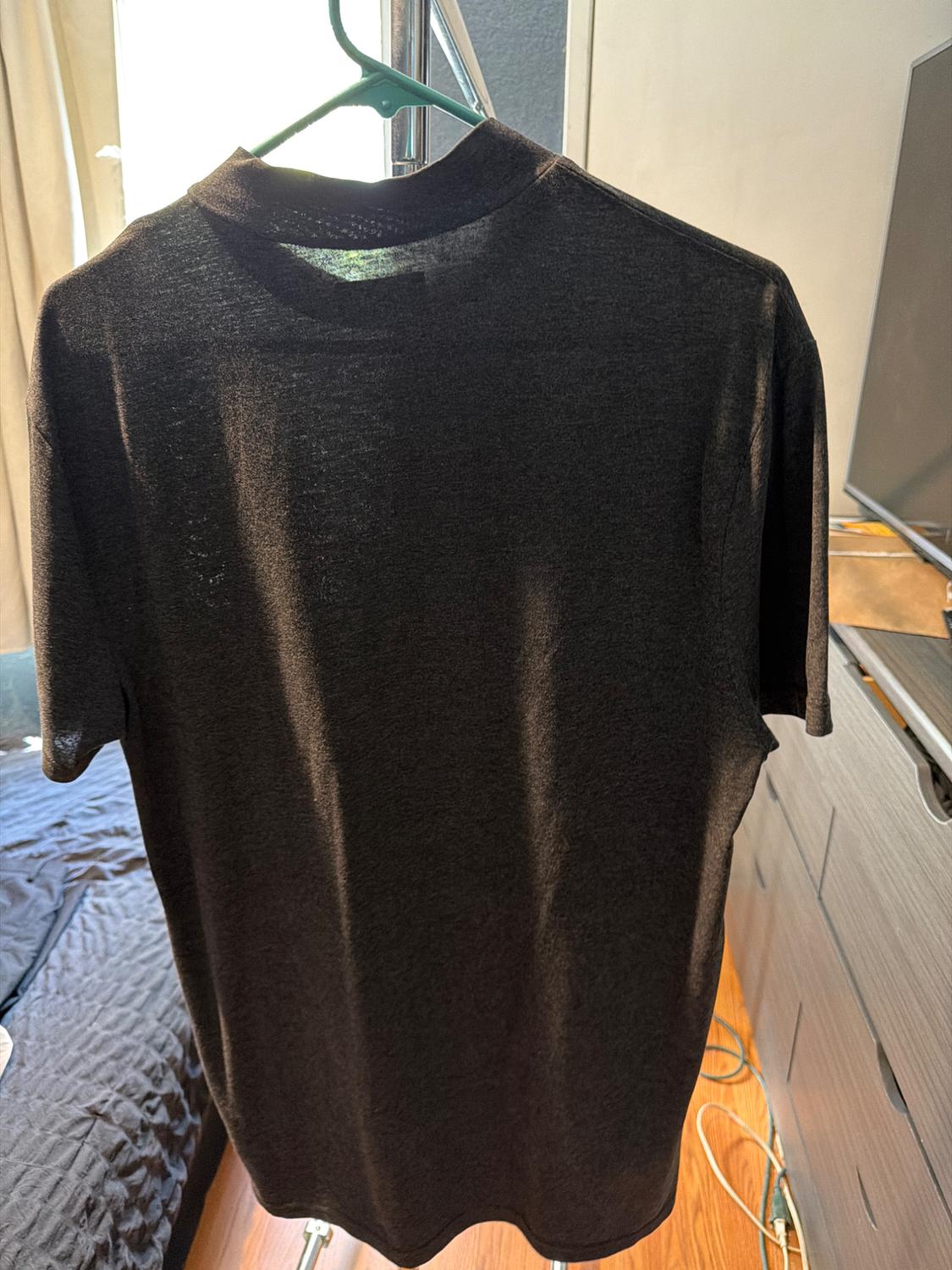 Dark gray short-sleeved t-shirt hanging on a green hanger in a sunlit room, with a bed and furniture in the background.