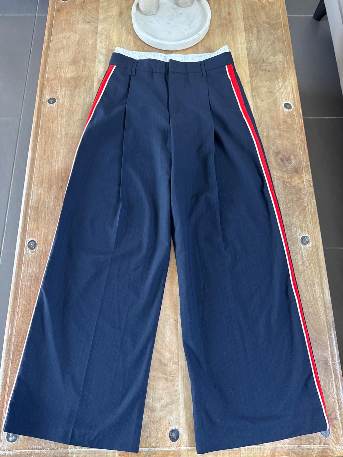 Navy blue wide-leg trousers with red and white stripes down the sides, on a light wooden table.
