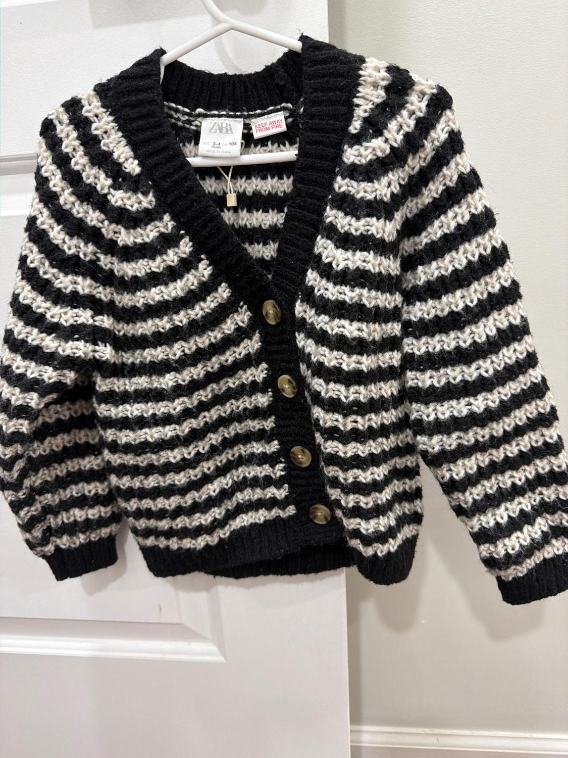 Black and white striped knit cardigan with brown buttons, hanging on a white hanger in front of a white door.