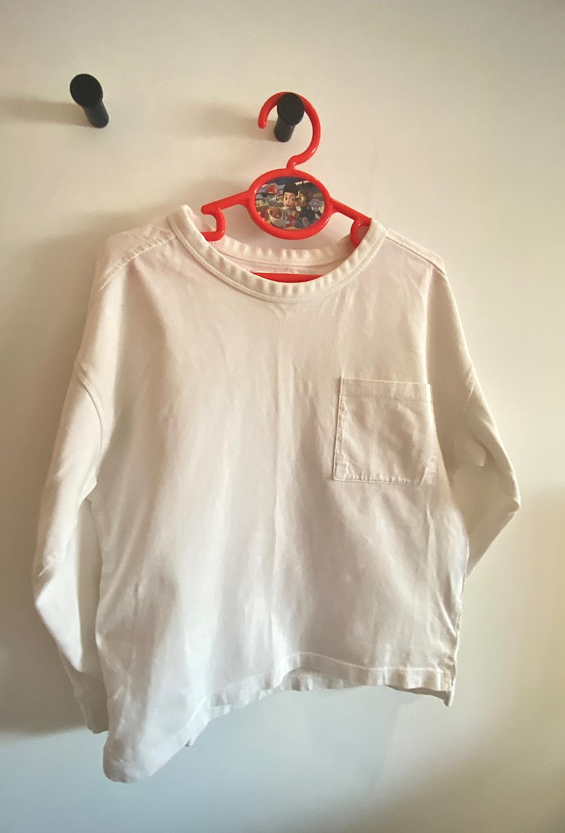 White long-sleeved T-shirt with a chest pocket, hanging on a red hanger with a children's design against a white wall.
