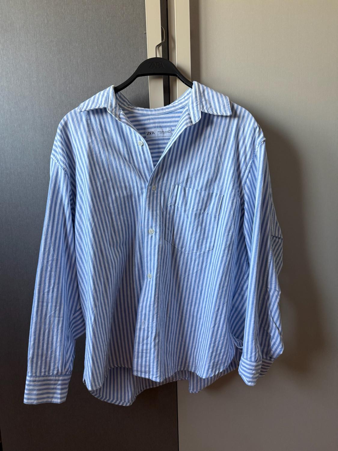 Blue and white striped shirt hanging on a black hanger. ZARA text on the hanger and label. Long-sleeved design with a chest pocket.