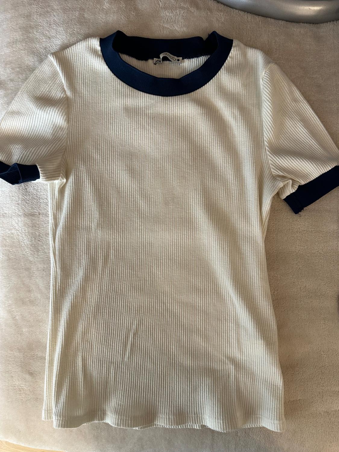 White ribbed T-shirt with navy blue collar and sleeves, on a beige surface.