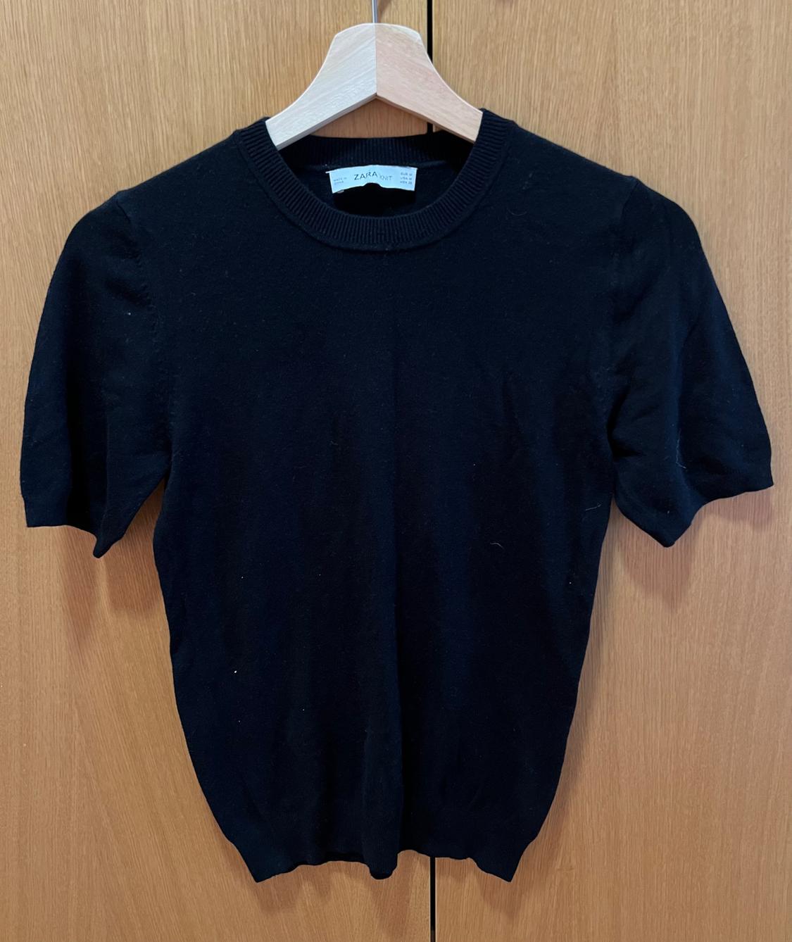 Black short-sleeved knitted top hanging on a wooden hanger against a wooden wardrobe background. Label with the text ZARA KNIT.