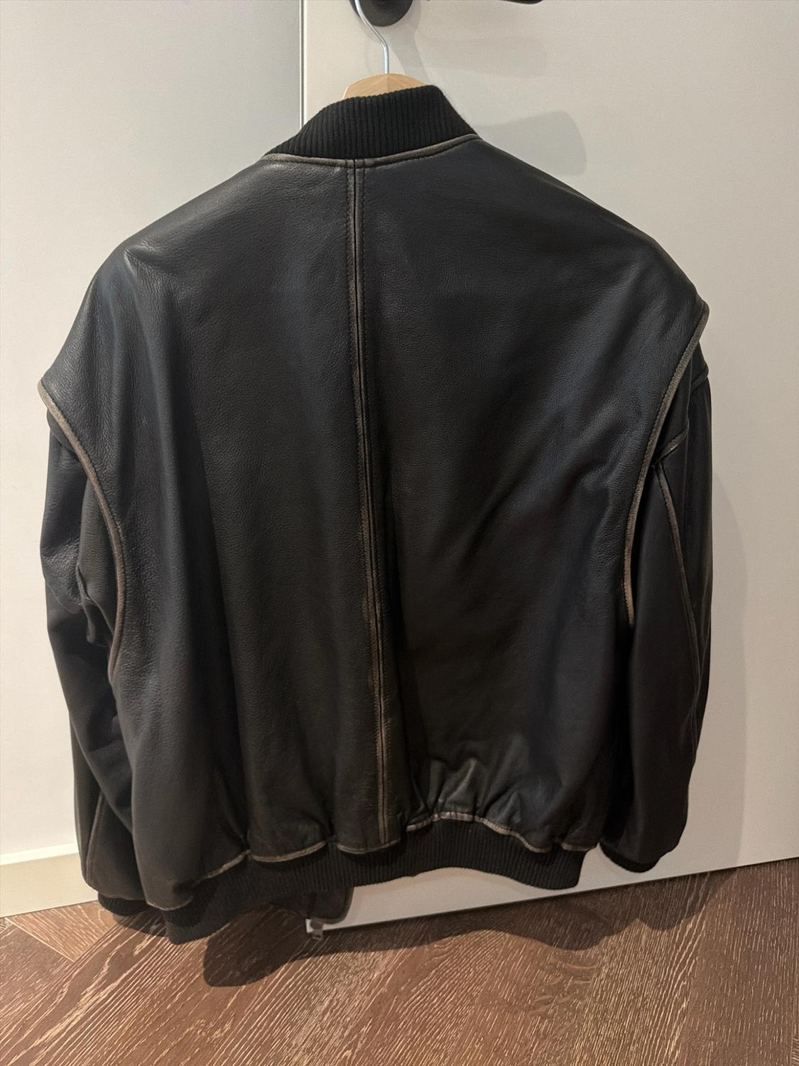 Rear view of a black leather jacket hanging on a white door. The ribbed collar and hem stand out alongside the stitching.