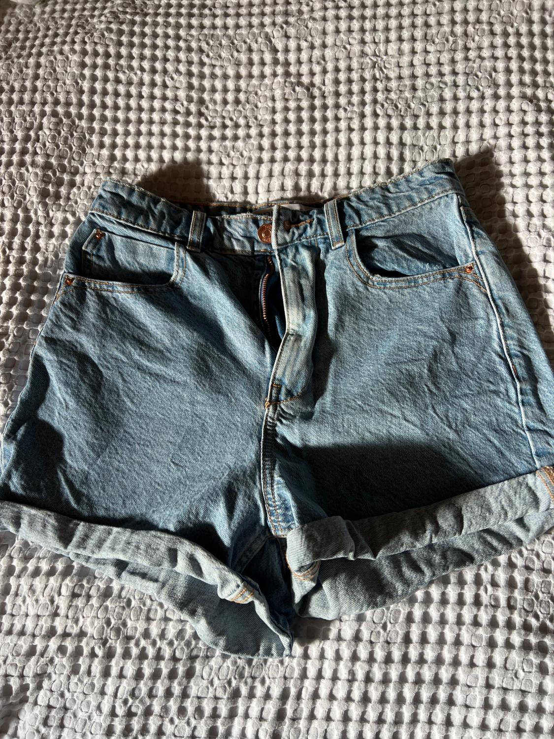 Unbuttoned denim shorts with a turned-up hem over textured white fabric. Natural light and sharp shadows create a relaxed style.
