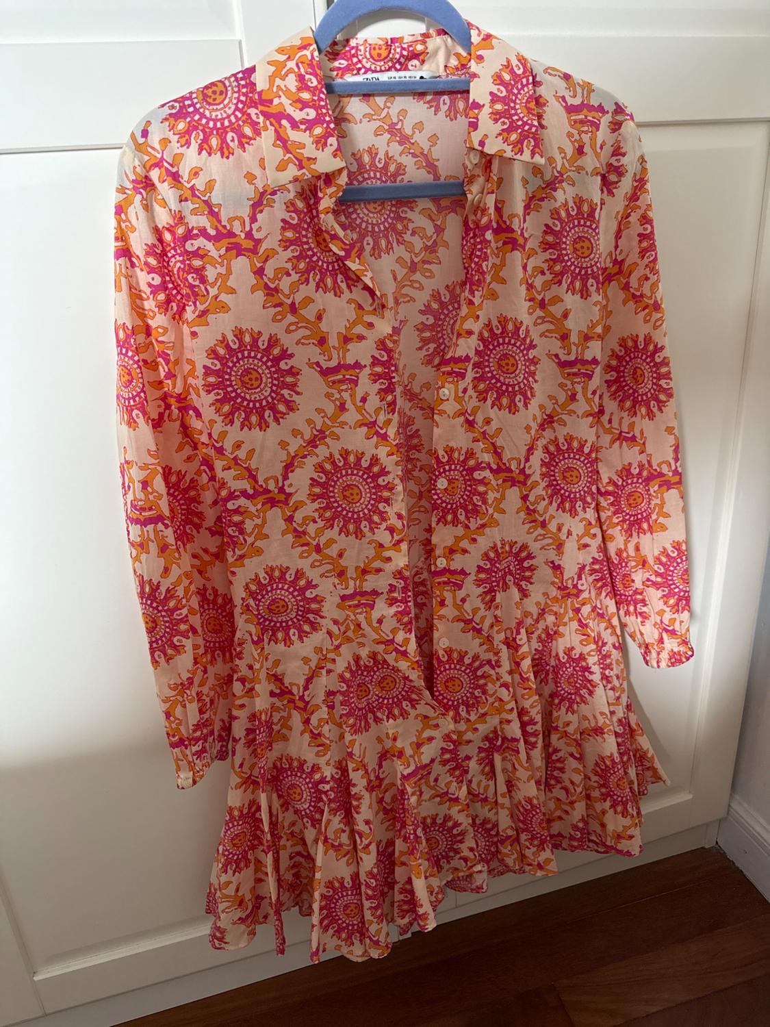 Long-sleeved shirt dress with a pink and orange circular print and a ruffled hem, hanging on a blue hanger over a white wardrobe.