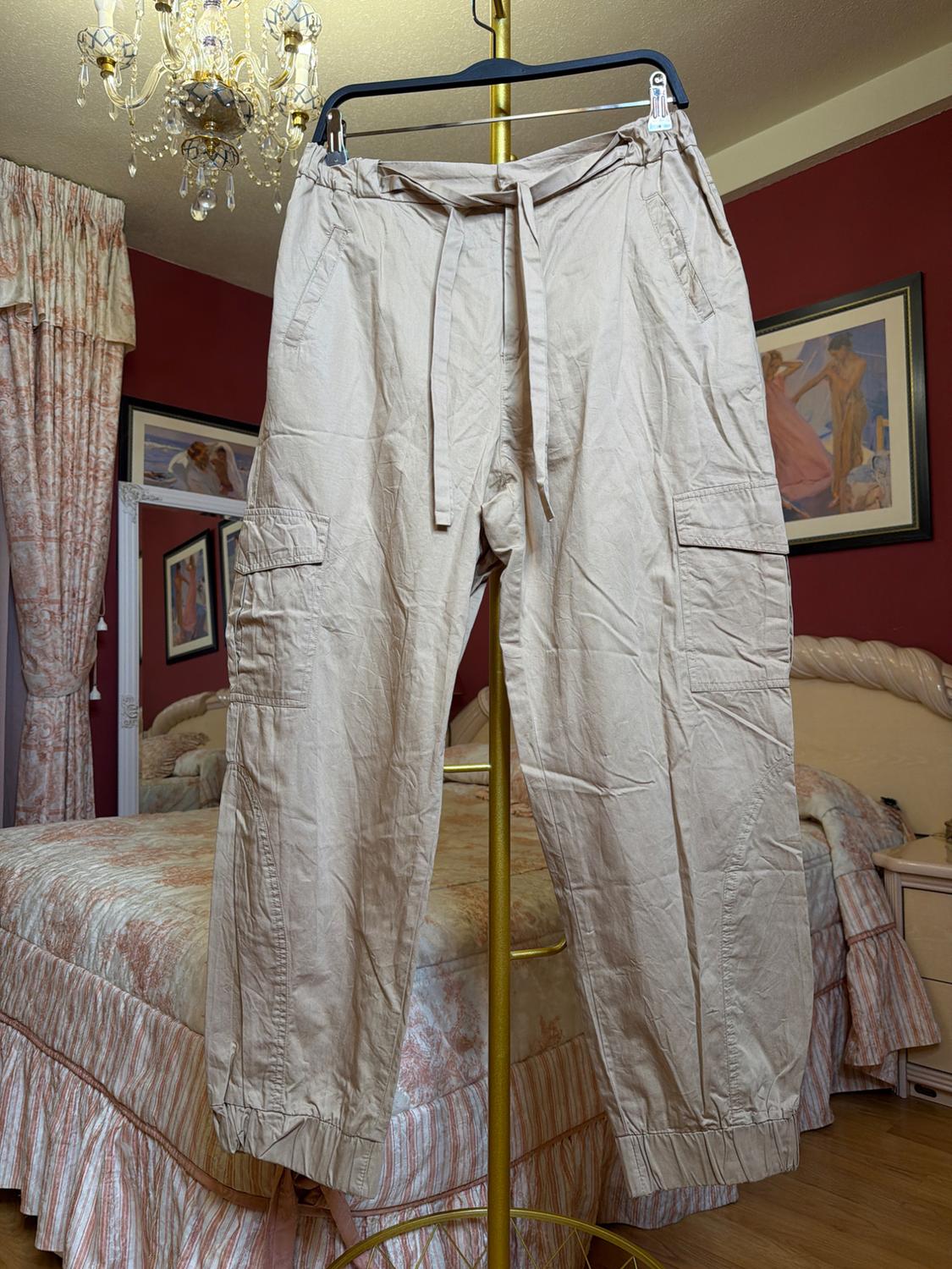 Beige trousers hanging on a hanger in a bedroom with curtains and pictures on the wall. Bed with a patterned bedspread.