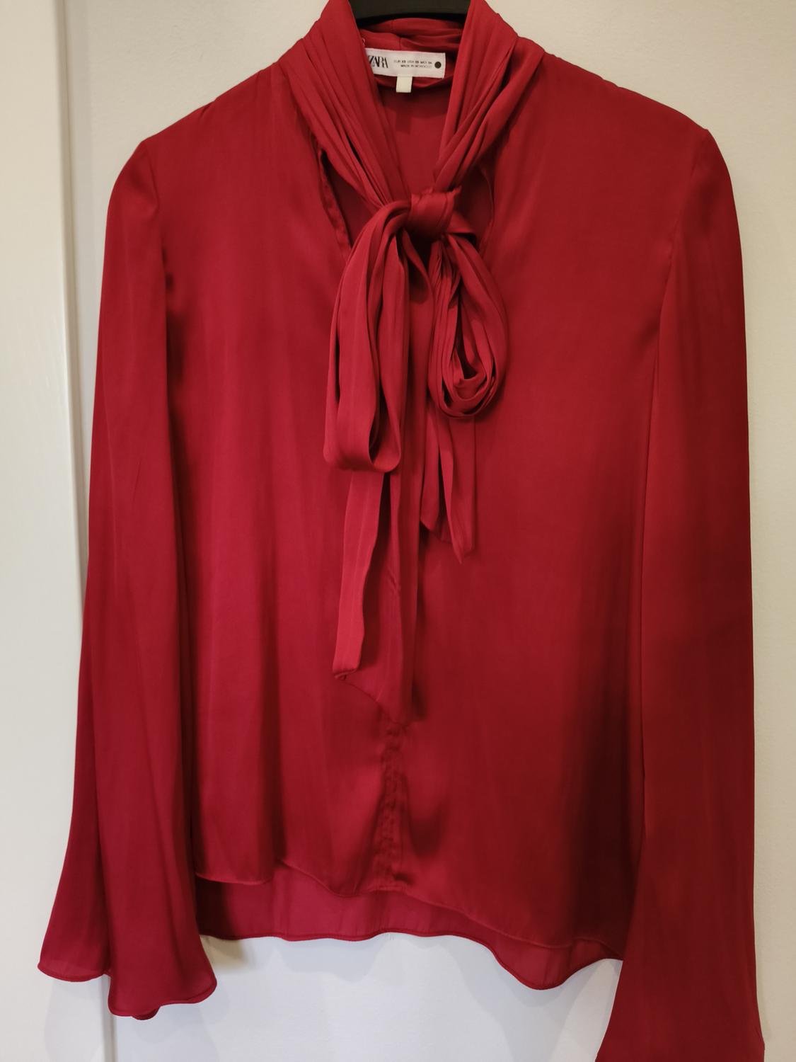 Red long-sleeved blouse with a bow at the neck, hanging on a hanger against a white background. Label visible inside.
