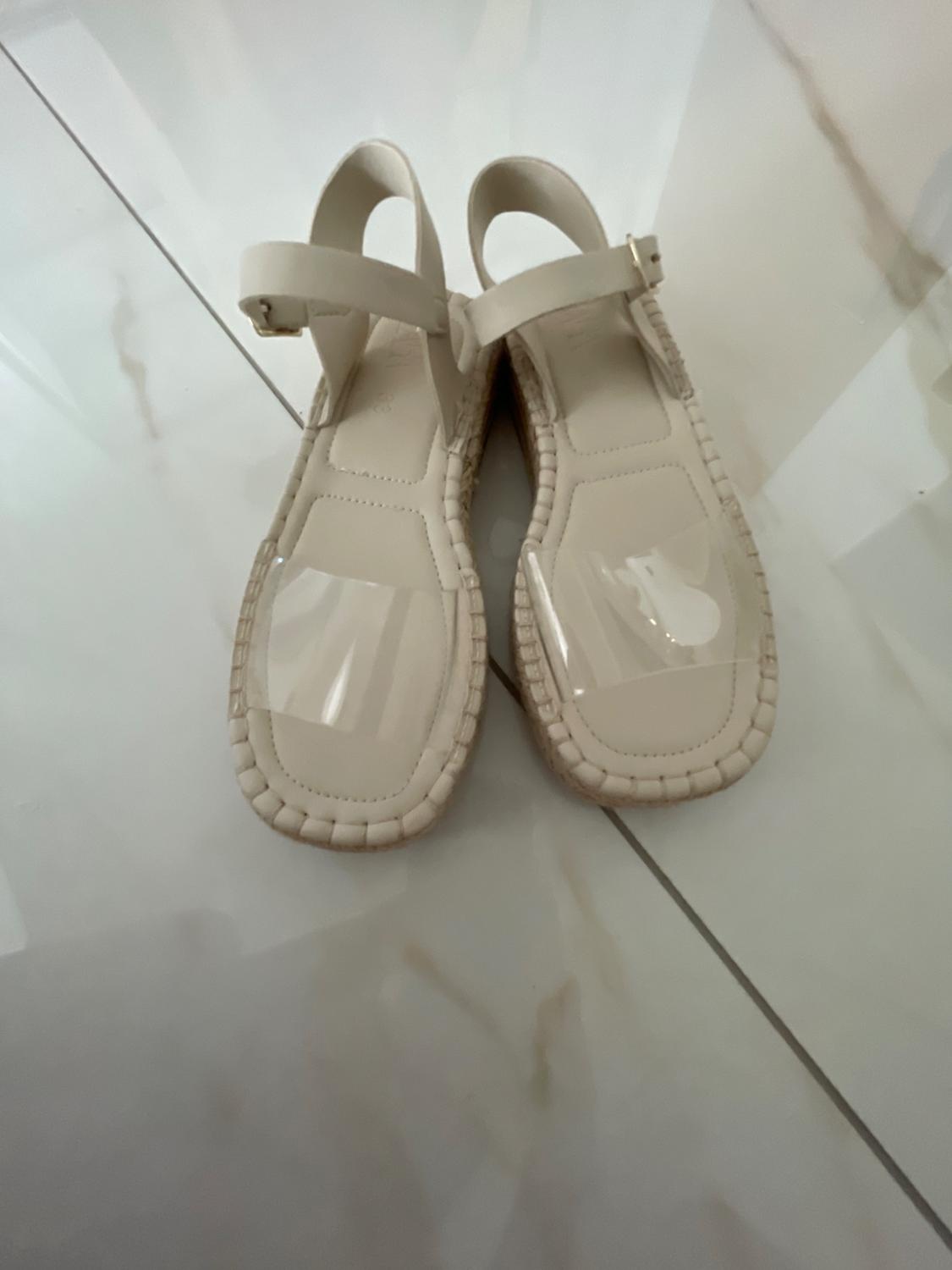 Cream-coloured sandals with jute soles, transparent front strap and ankle strap fastening with a buckle, on a light-tiled floor.