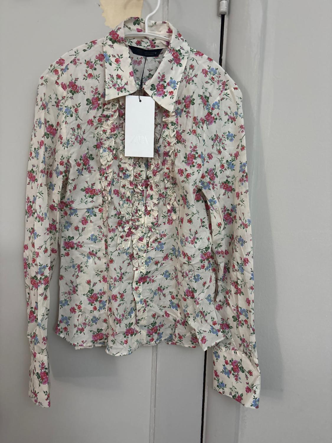Pink and blue floral-print blouse with a collar and front frills. Hanging on a hanger over a door with the ZARA label.