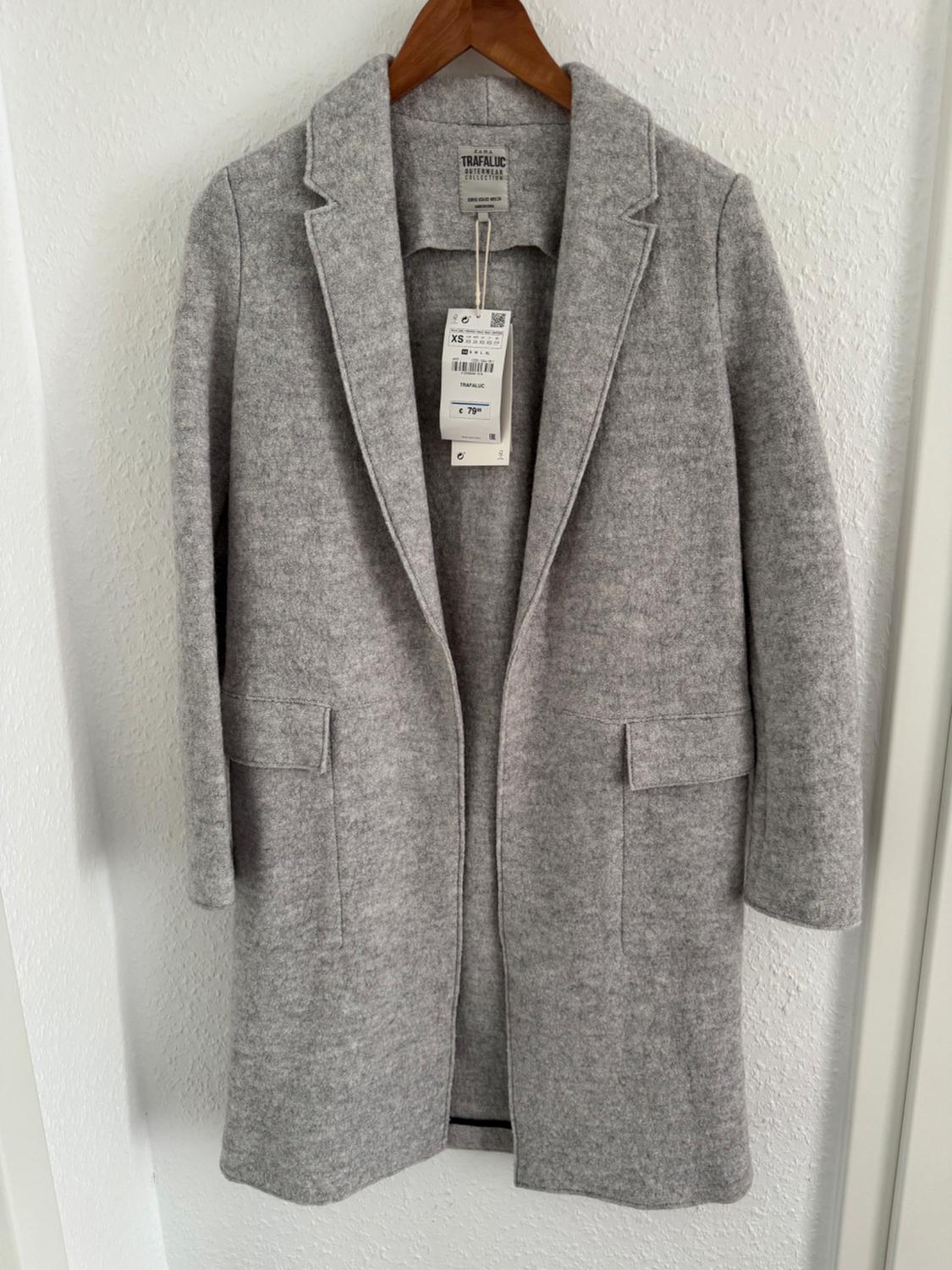 Light grey coat hanging on a wooden hanger. Visible tag with a price of 79 euros and size XS. White wall background.