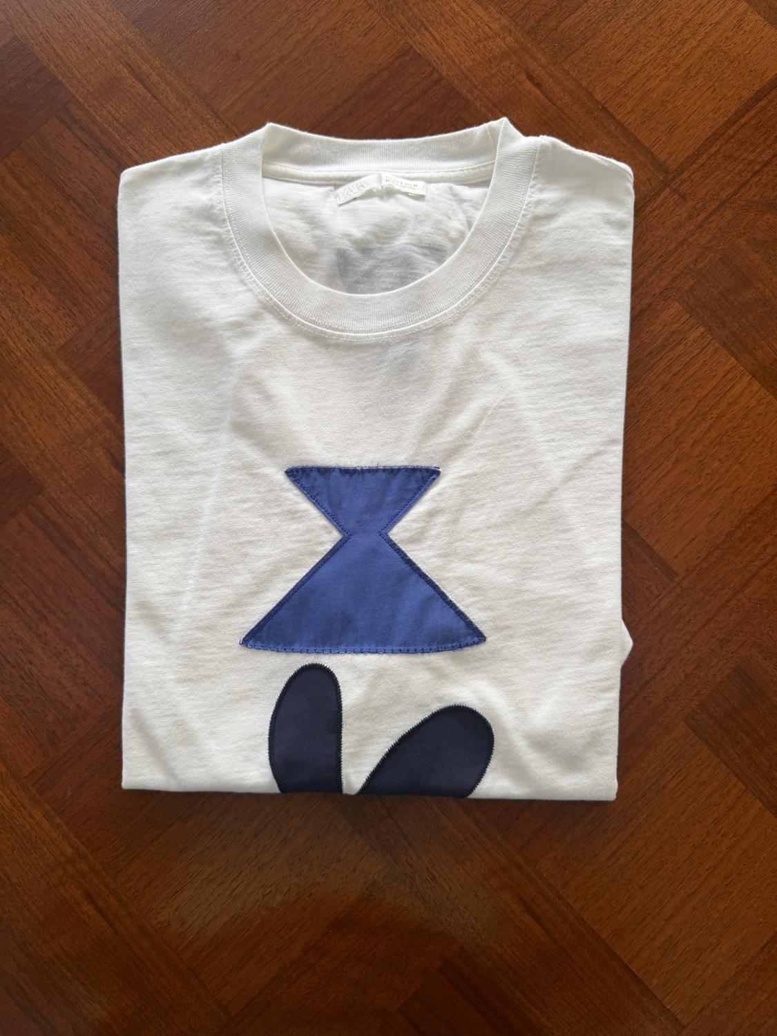 Λευκό διπλωμένο T-shirt σε ξύλινο πάτωμα. Ξεχωρίζει ένα σχέδιο στο μπροστινό μέρος με μια μπλε γεωμετρική φιγούρα και δύο σκούρα οβάλ σχήματα.