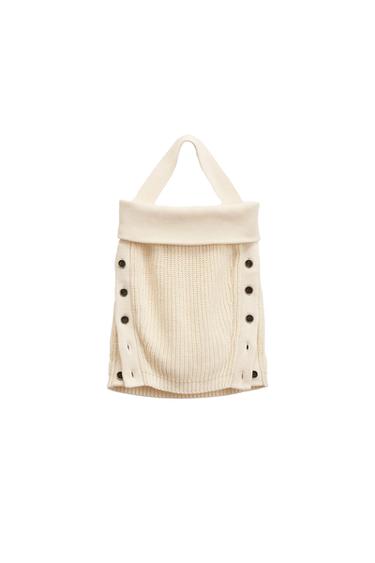 Cream knit bag with black handles and buttons on the sides, on a white background.