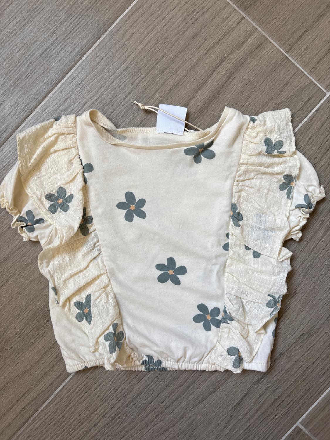 Girl's short-sleeved blouse in a cream colour with a grey floral print and frill detailing, on a grey tiled background.