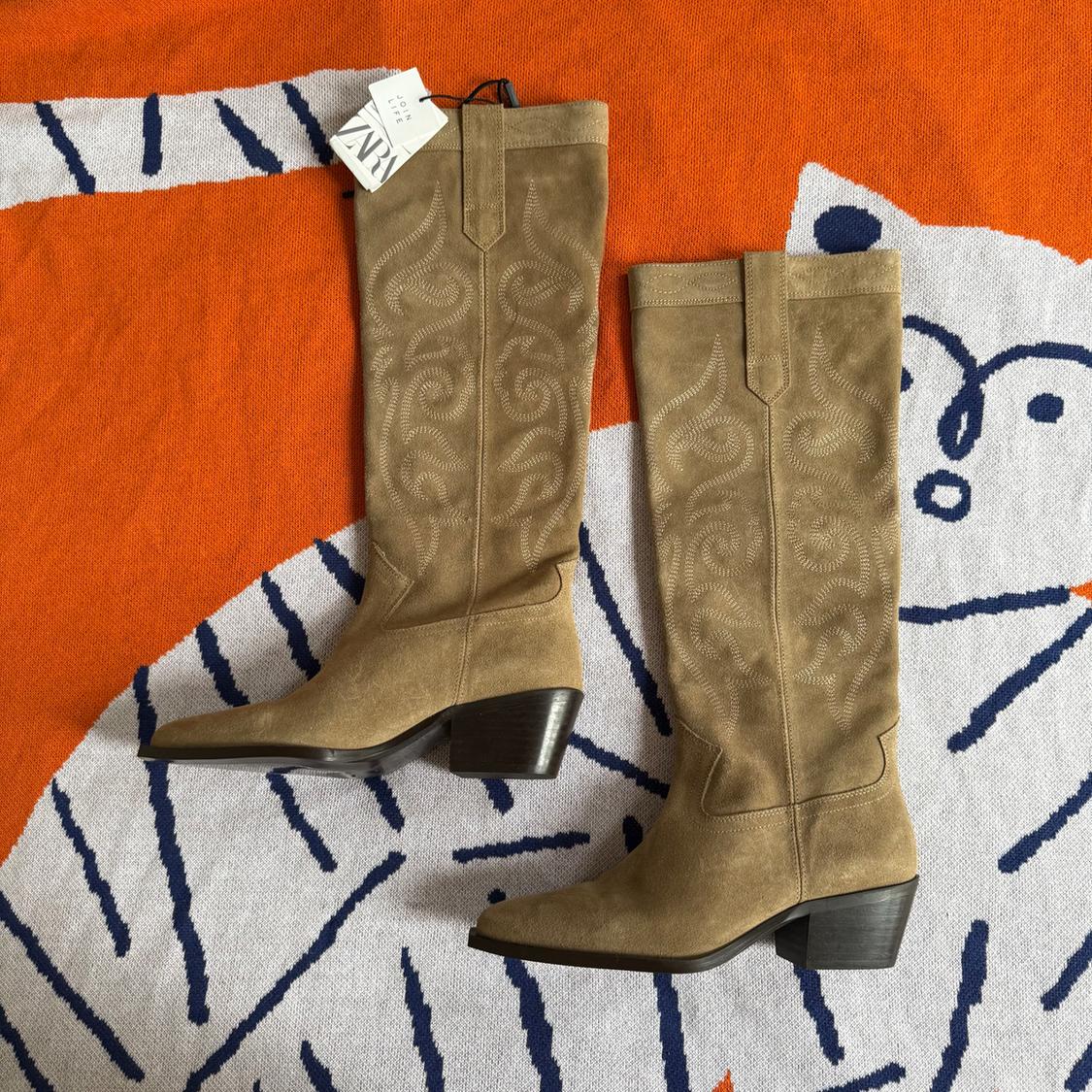 Beige suede high-heeled boots with a chunky heel on an orange blanket with a blue and white tiger design. Visible tag.