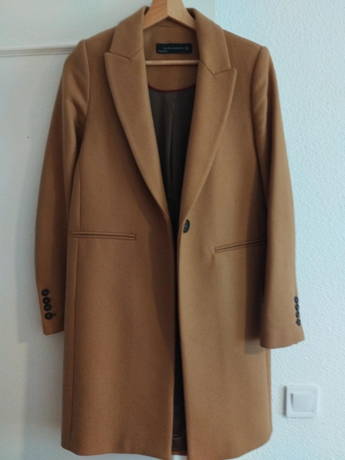Camel coat hanging on a wooden hanger, with wide lapels and buttons on the sleeves. Plain white wall background.