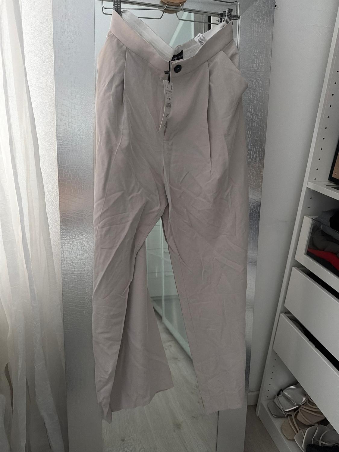 Creased beige trousers hanging on a hanger in front of a mirror. Background with white curtains and a shoe rack.